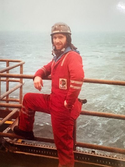 A young Jimi Gill in red offshore coveralls and hard hat at the handrail, grey sea behind.