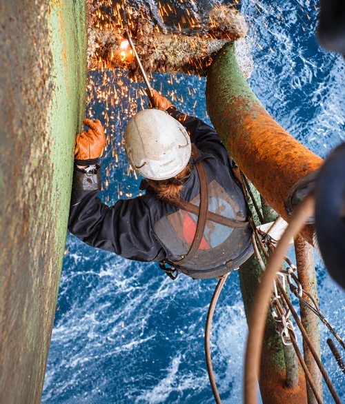 A rope access technician cutting steel above the sea, sparks falling.