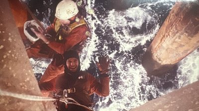 Two rope access technicians suspended over breaking sea beneath a platform.